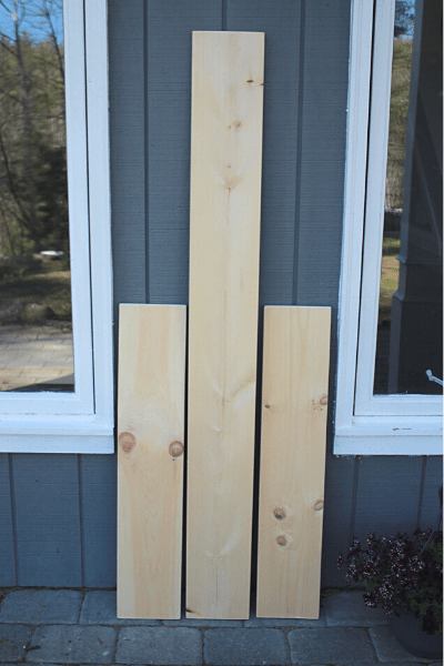 one long piece of wood and two shorter pieces leaning up against a grey house with windows on each side