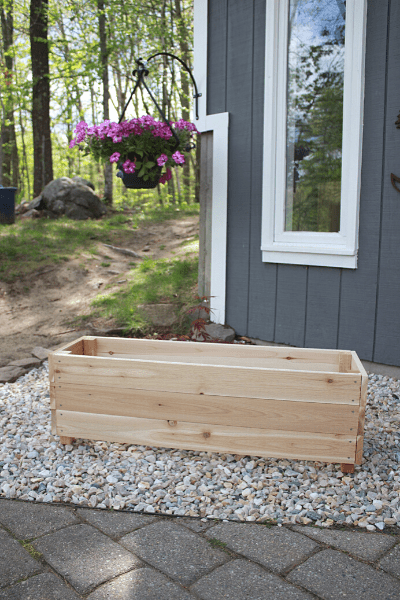 DIY planter box sitting on top of river rocks next to a grey house with purple flowers hanging behind 