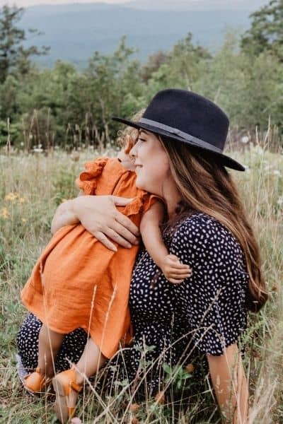 mother wearing a black hat sitting on the ground in a field with her toddler hugging her