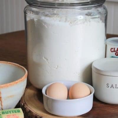 two eggs in a small bowl sitting next to a salt jar, a stick of butter, and a large glass anchor jar filled with flour