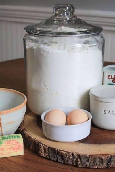 two eggs in a small bowl sitting next to a salt jar, a stick of butter, and a large glass anchor jar filled with flour